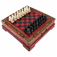 Échiquier de 26 x 26 cm rouge et noir sur socle à pieds, décoré de motifs dorés peints à la main, avec 32 pièces d’échecs figuratives en résine crème et noire.